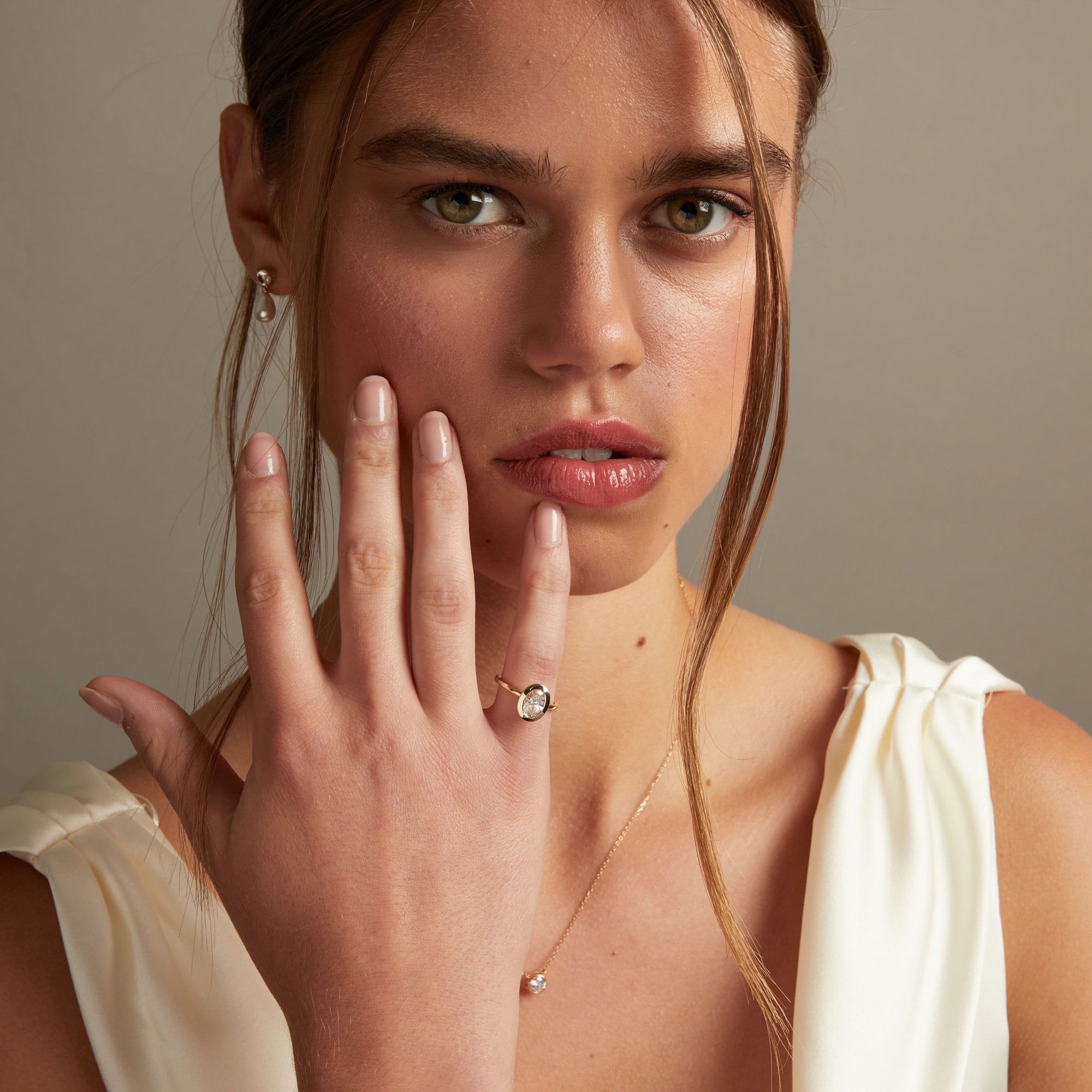 Model. A close-up lifestyle shot of a woman wearing a delicate gold ring with an oval clear diamond on her finger, paired with a matching gold necklace. The background is a soft, neutral gray.