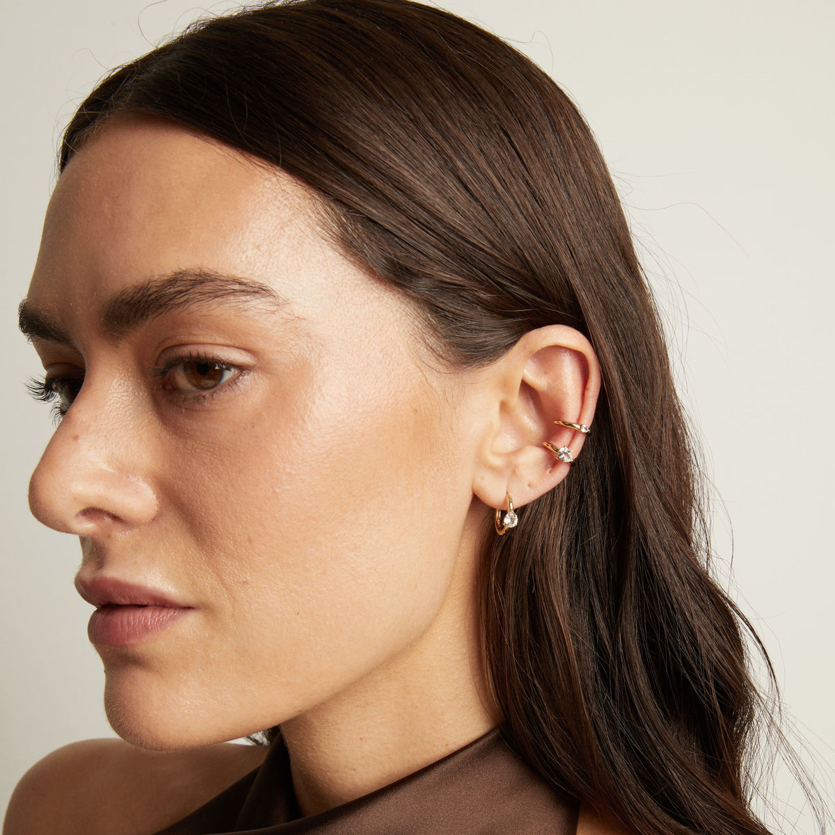 Close up of woman wearing two gold ear cuffs and a huggie earring with white sapphires on neutral background. 