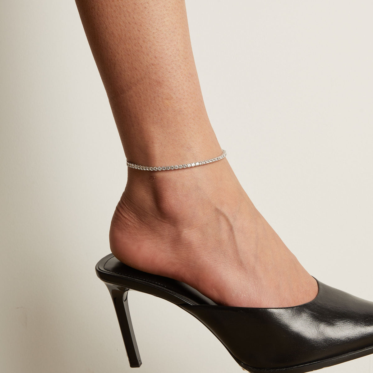 Model. Tennis anklet and black high heeled shoe worn on a foot with a neutral background.