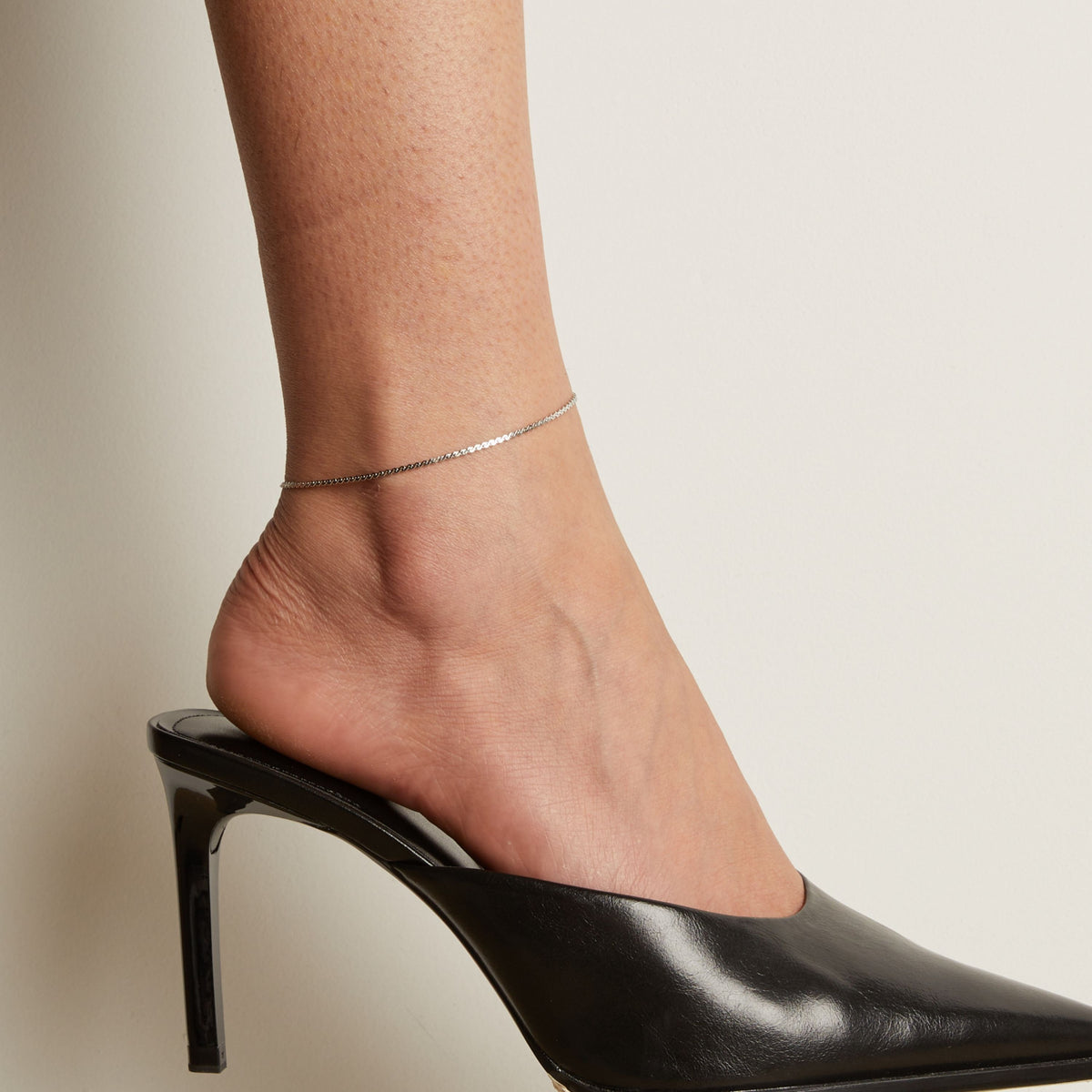 Silver anklet on a foot wearing a black high heeled shoe against a neutral background.