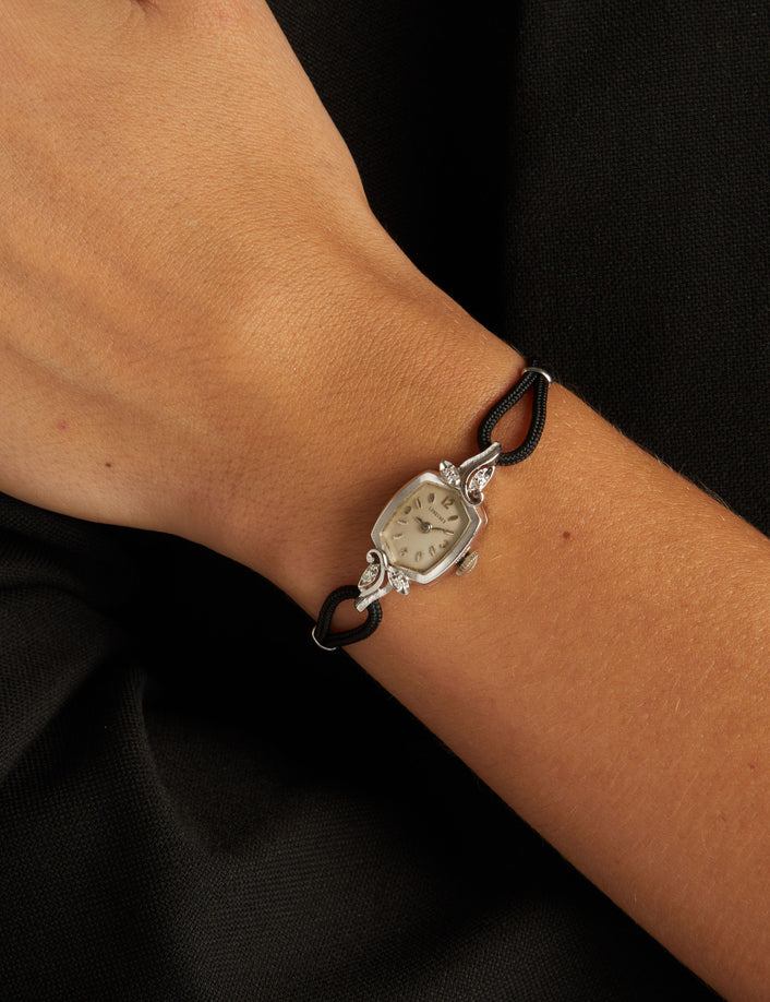Model. Close up of a woman wearing a black rope watch with silver face with diamonds on neutral background.
