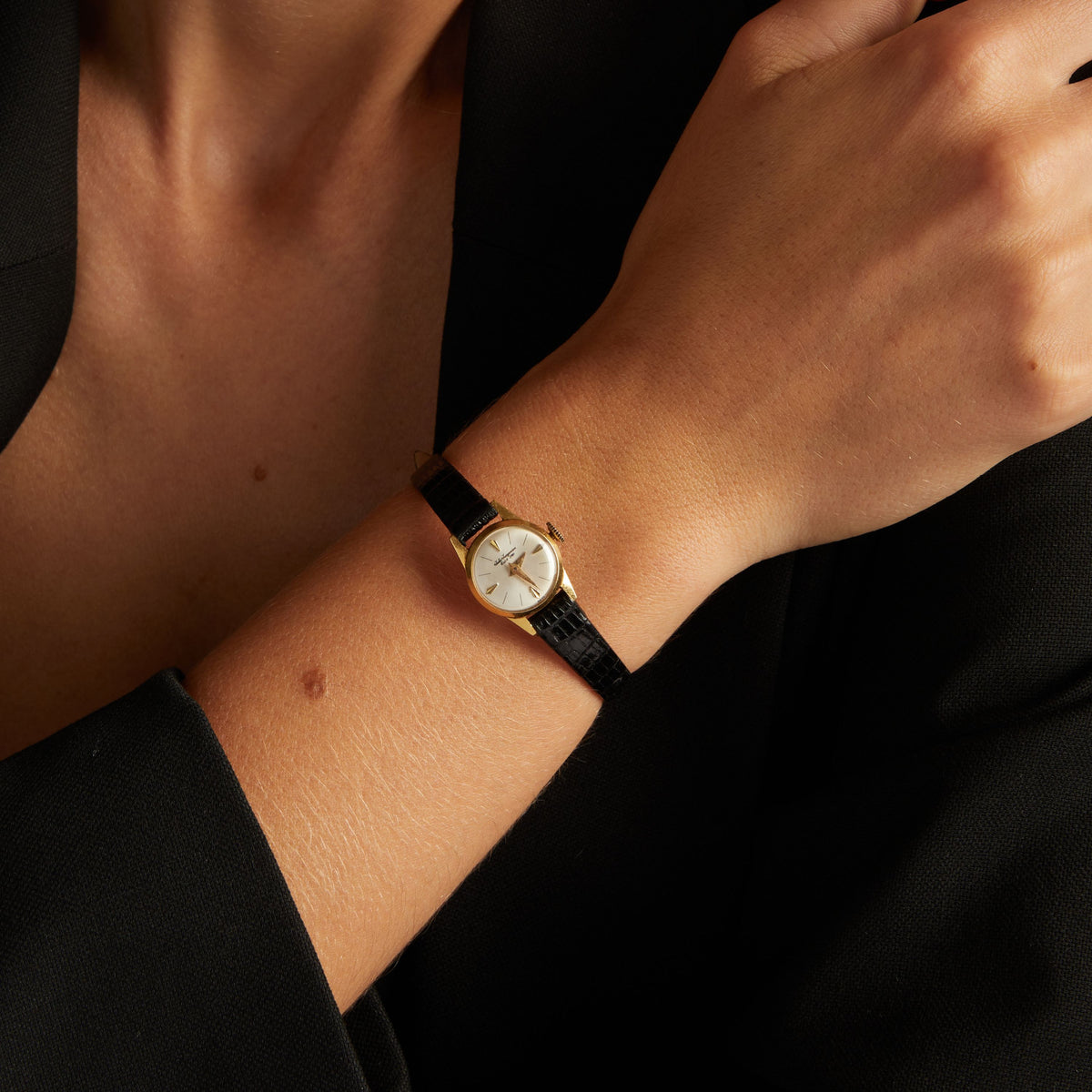 Model. Close up of a woman wearing a black leather watch with gold face on neutral background.