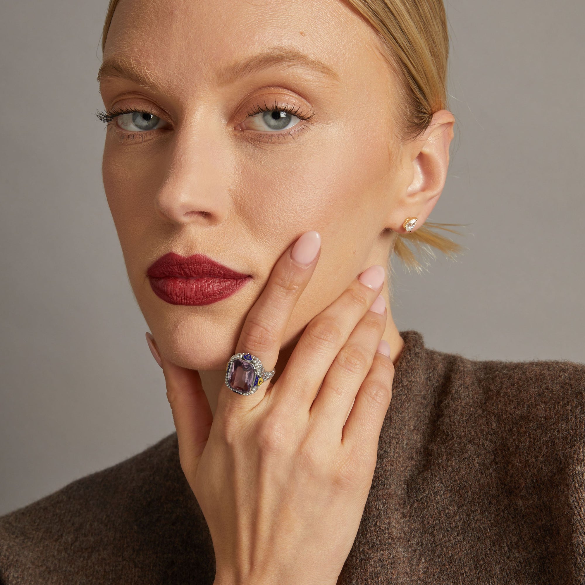 Model. Close up of woman wearing bold purple gemstone ring on neutral background.