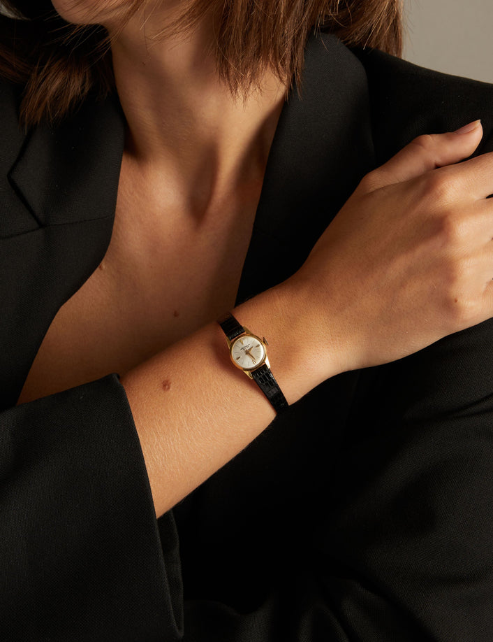 Model. Close up of a woman wearing a black leather watch with gold face on neutral background.