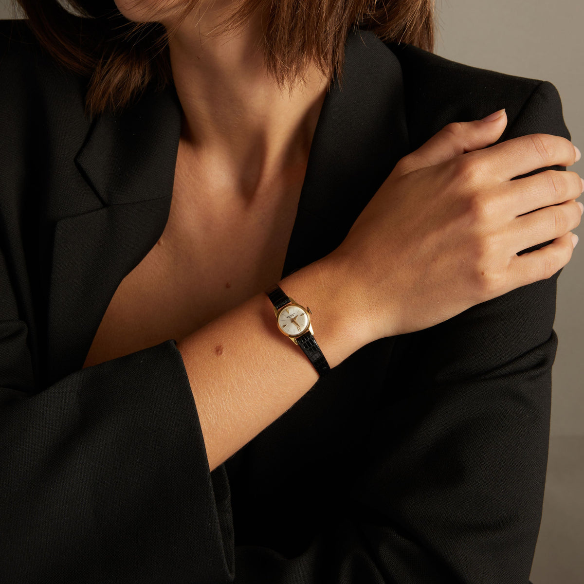 Model. Close up of a woman wearing a black leather watch with gold face on neutral background.