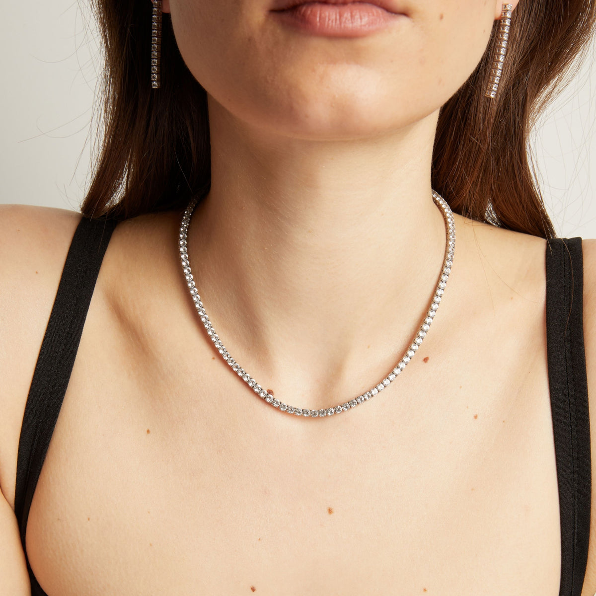 Close up of a woman wearing a tennis necklace and earrings against a neutral background.