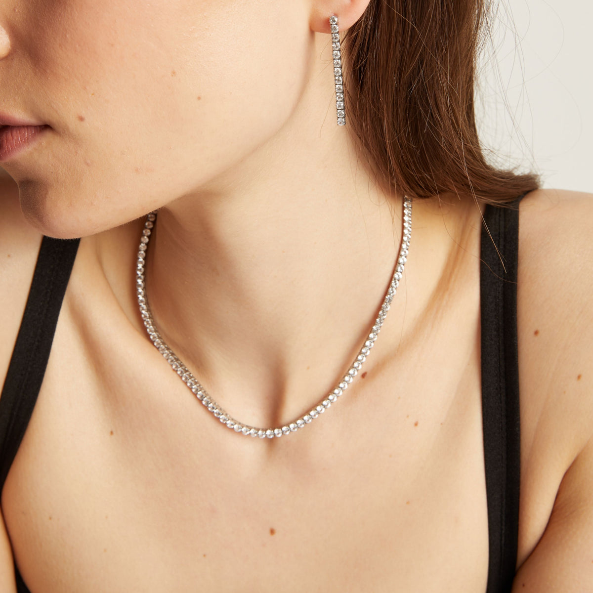 Close up of a woman wearing a tennis necklace and earrings against a neutral background.