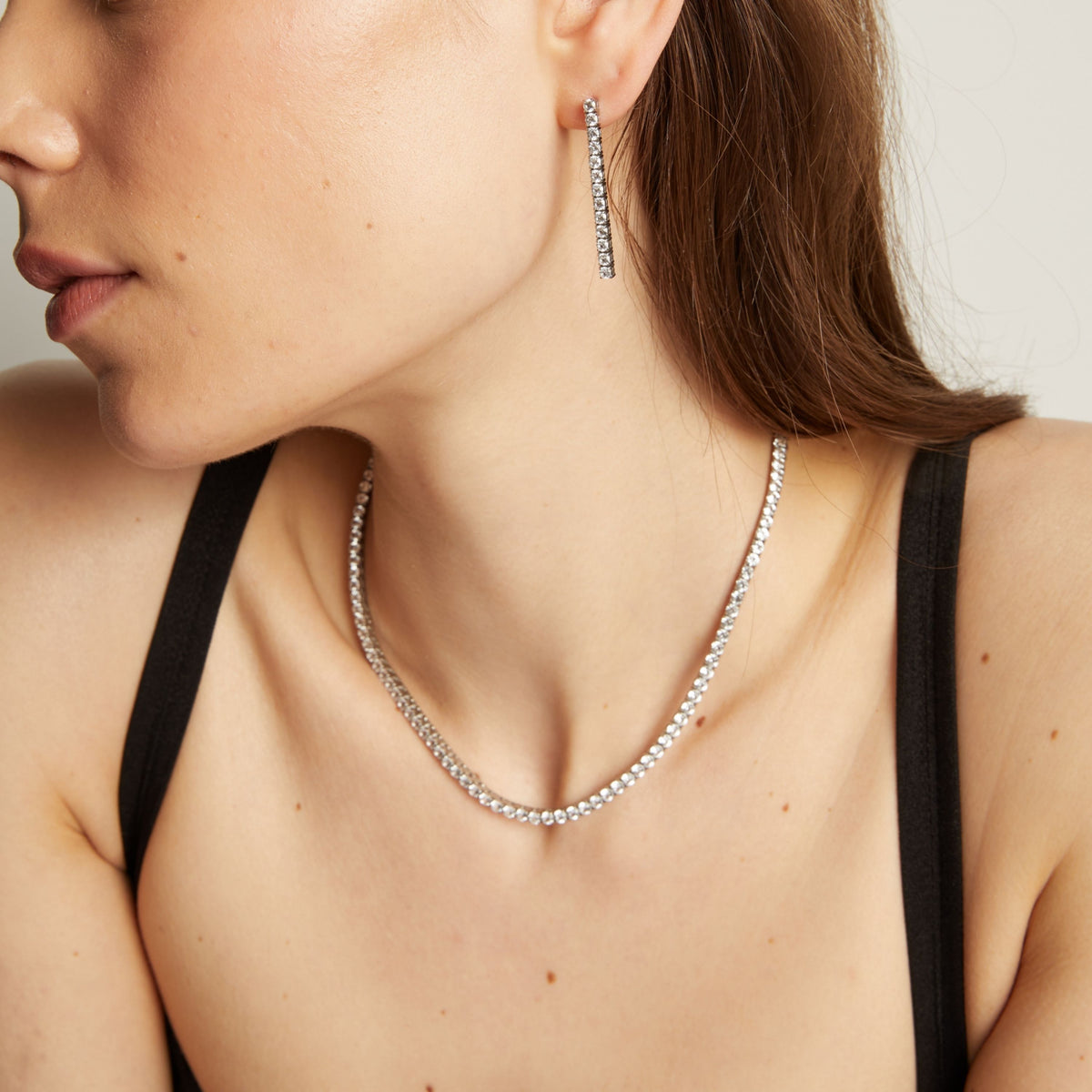Close up of a woman wearing a tennis necklace and earrings against a neutral background.