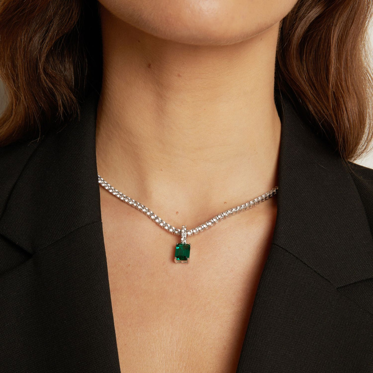 Model. Close up of a woman wearing a black blazer with a tennis necklace with a emerald gemstone pendant.