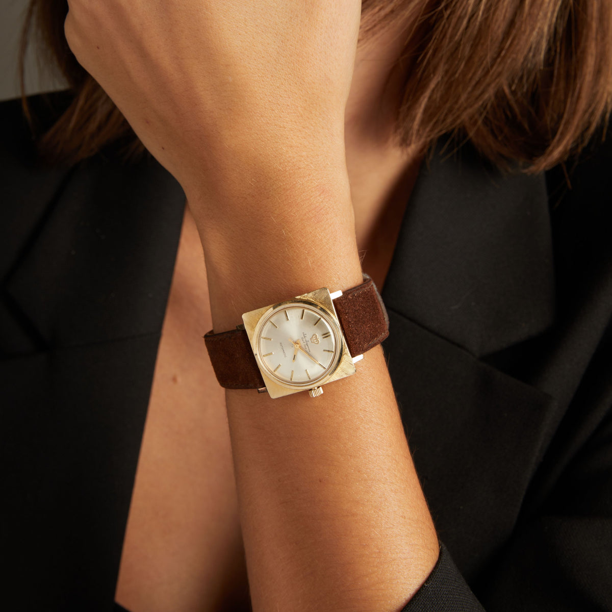 Model. Close up of a woman wearing a brown leather watch with gold face on neutral background.