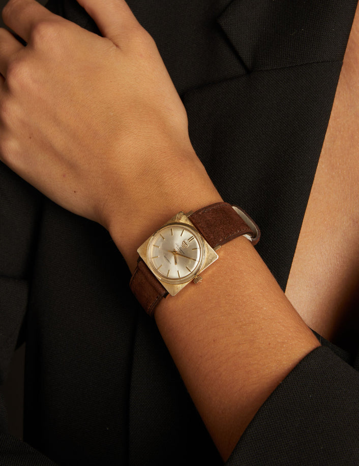 Model. Close up of a woman wearing a brown leather watch with gold face on neutral background.