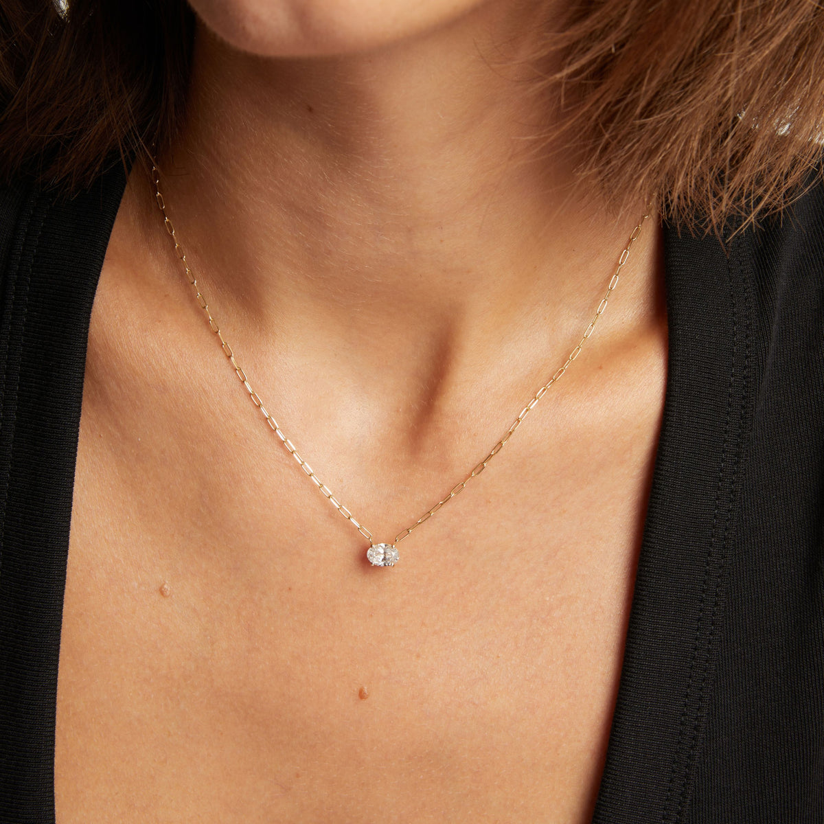 Close up of a woman in a black top wearing a chain necklace with a solitaire oval diamond on neutral background.
