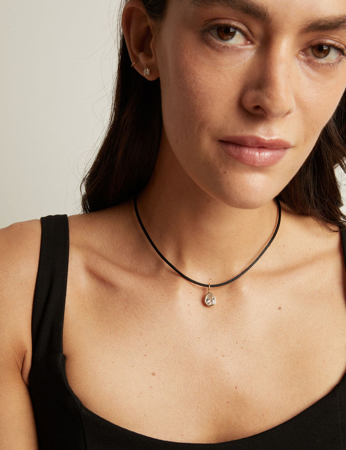Model. A close-up of a woman wearing a delicate black cord necklace with a small silver-toned pendant featuring a clear teardrop-shaped stone.