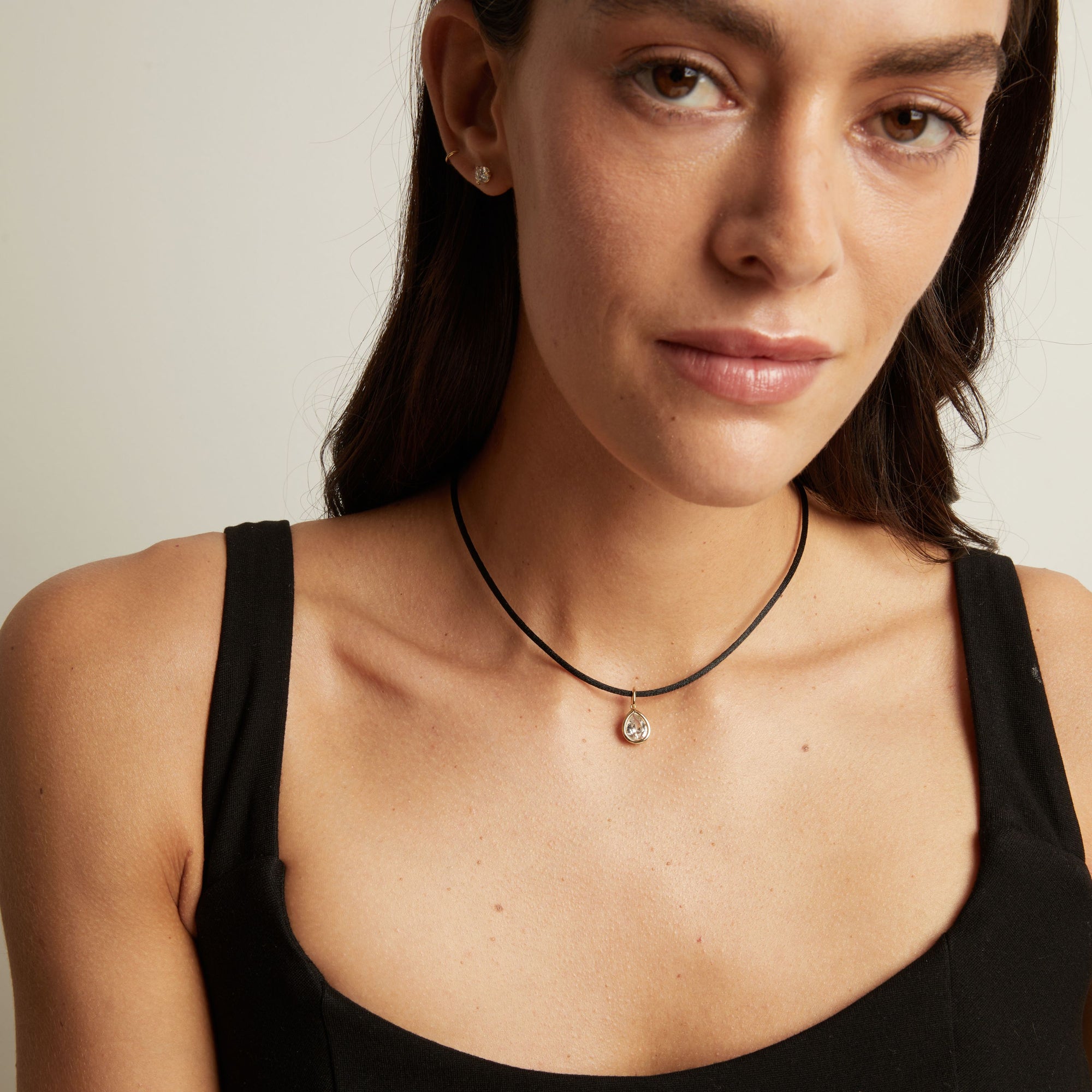 Model. A close-up of a woman wearing a delicate black cord necklace with a small silver-toned pendant featuring a clear teardrop-shaped stone.