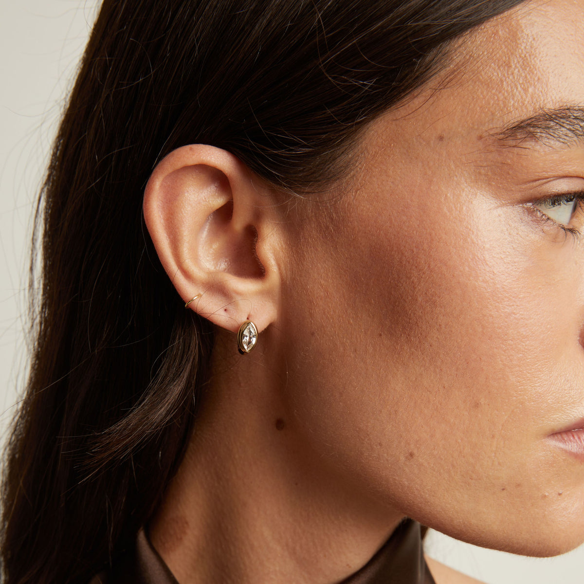 Close up of woman wearing a gold bezel huggie earring on neutral background.