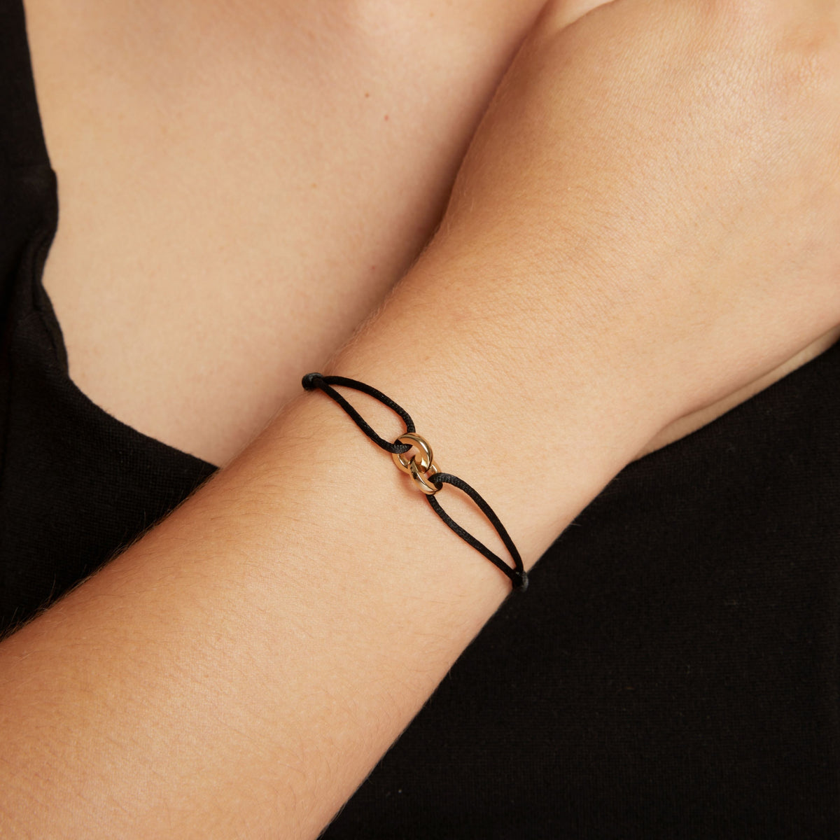 Model. A close-up of a person's wrist wearing a delicate cord bracelet with a small gold-toned interlocking link charm.