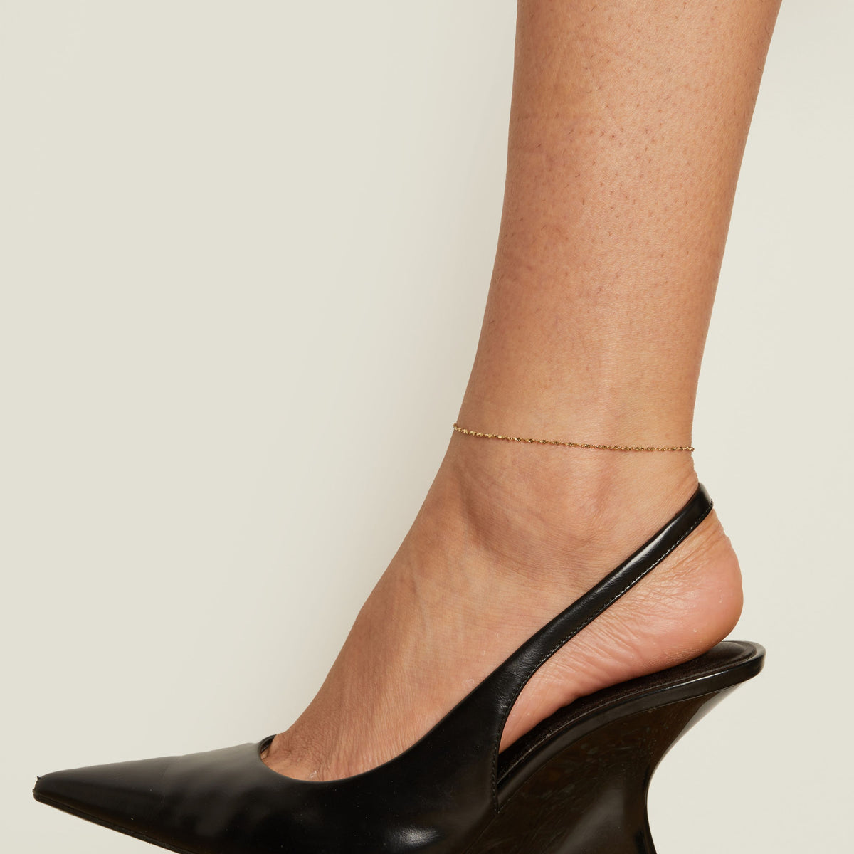 Black wedge heel shoe and anklet worn by a person on a neutral background