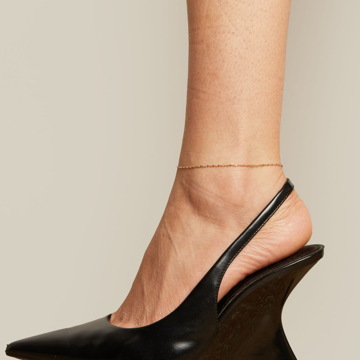 Model. Black wedge heel shoe and anklet worn by a person on a neutral background