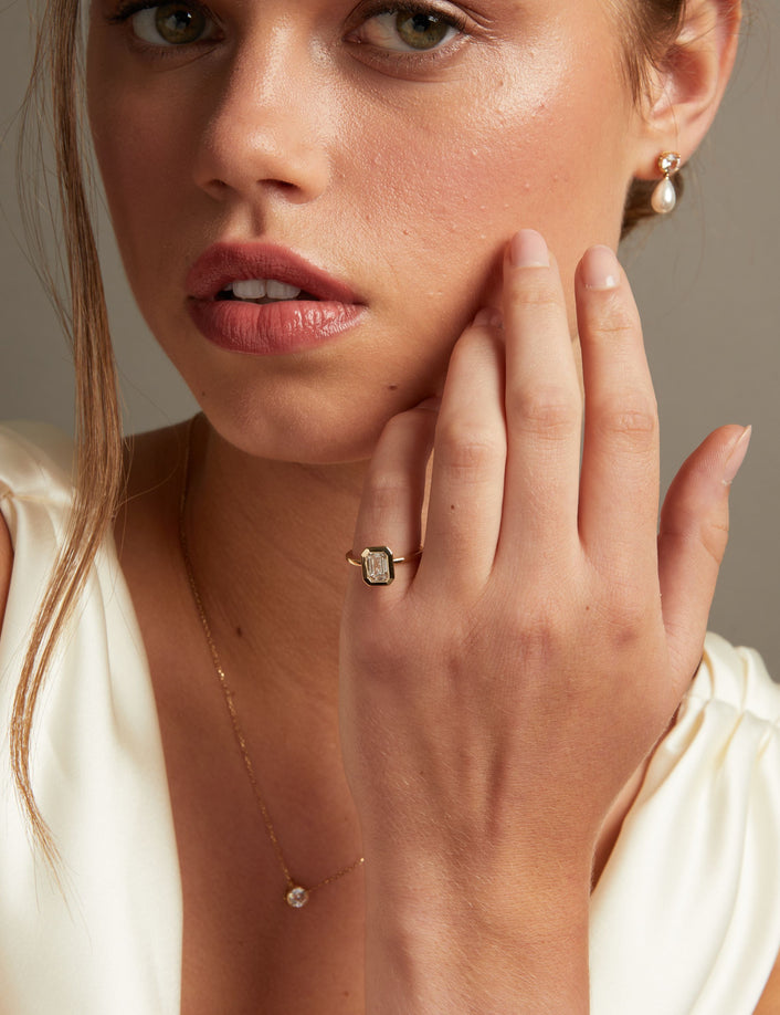 A close-up lifestyle shot of a woman wearing a gold ring with an emerald diamond on her finger, along with matching pearl drop earrings and a delicate gold necklace.