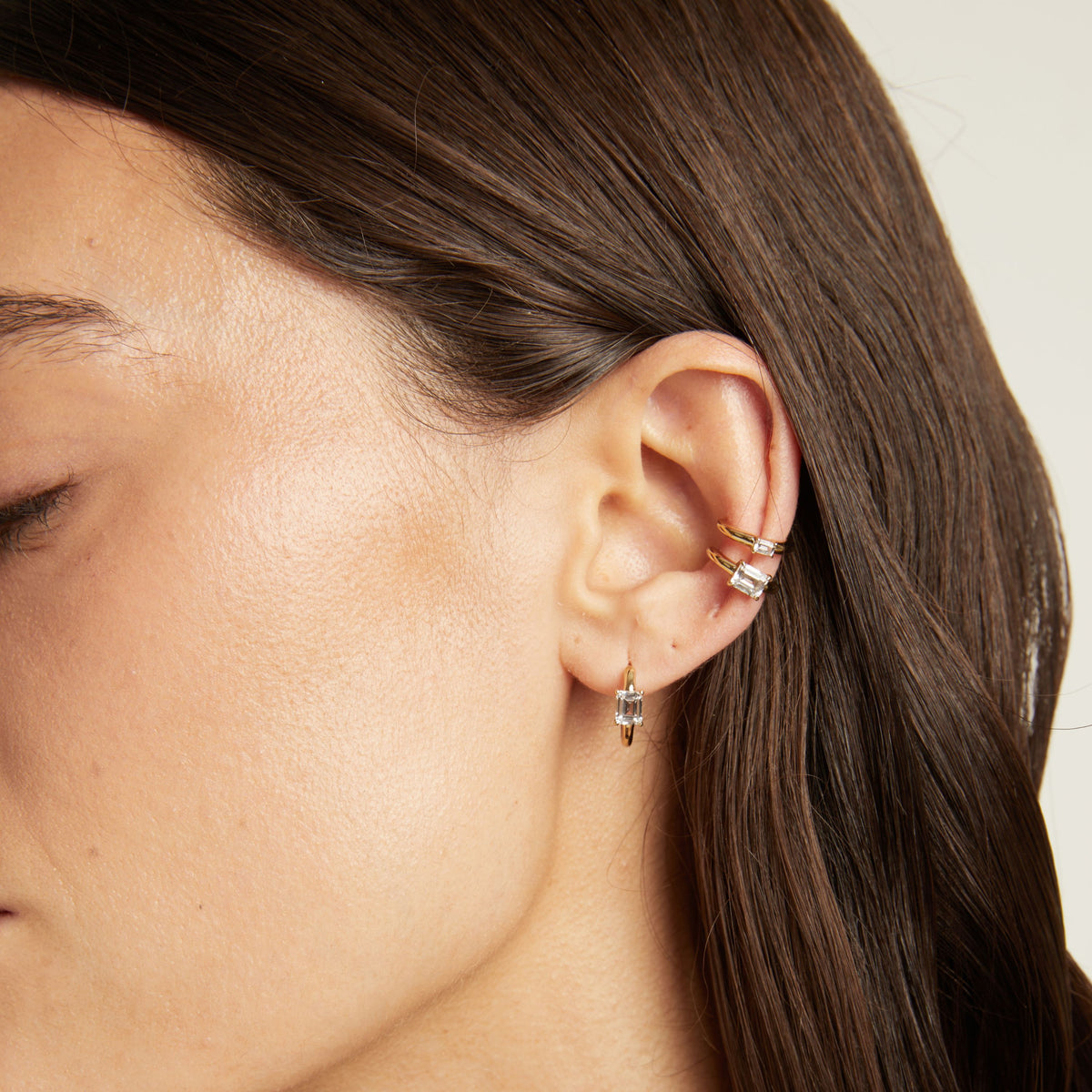 Close up of woman wearing two gold ear cuffs and a huggie earring with white sapphires on neutral background. 