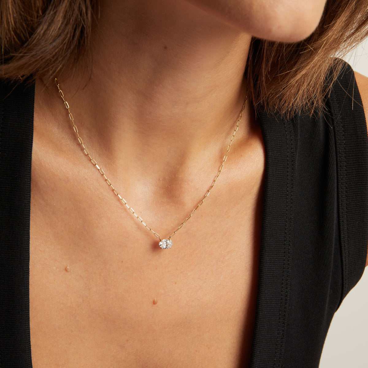 Close up of a woman in a black top wearing a chain necklace with a solitaire oval diamond on neutral background.