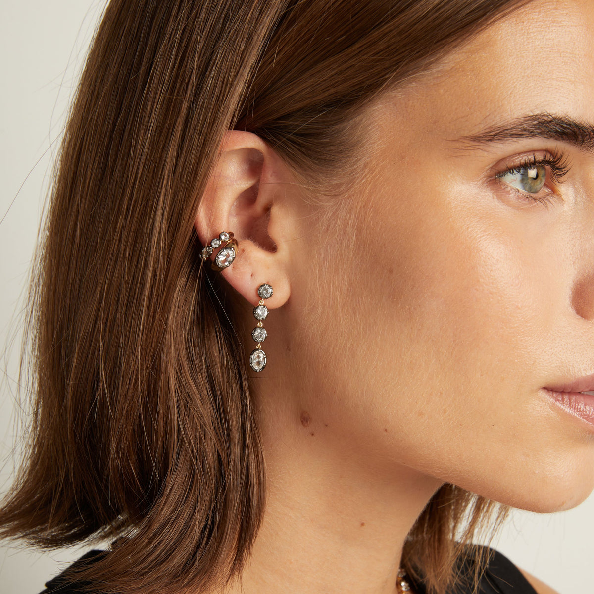 Model. Close up of woman wearing tennis earrings with two ear cuffs on neutral background.