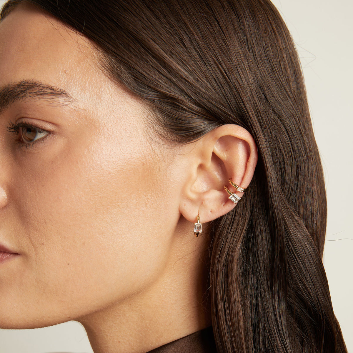 Model. Close up of woman wearing two gold ear cuffs and a huggie earring with white sapphires on neutral background. 