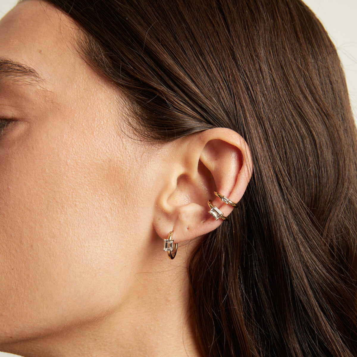 Close up of woman wearing two gold ear cuffs and a huggie earring with white sapphires on neutral background. 