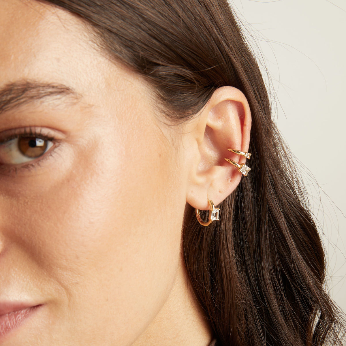 Close up of woman wearing two gold ear cuffs and a huggie earring with white sapphires on neutral background. 
