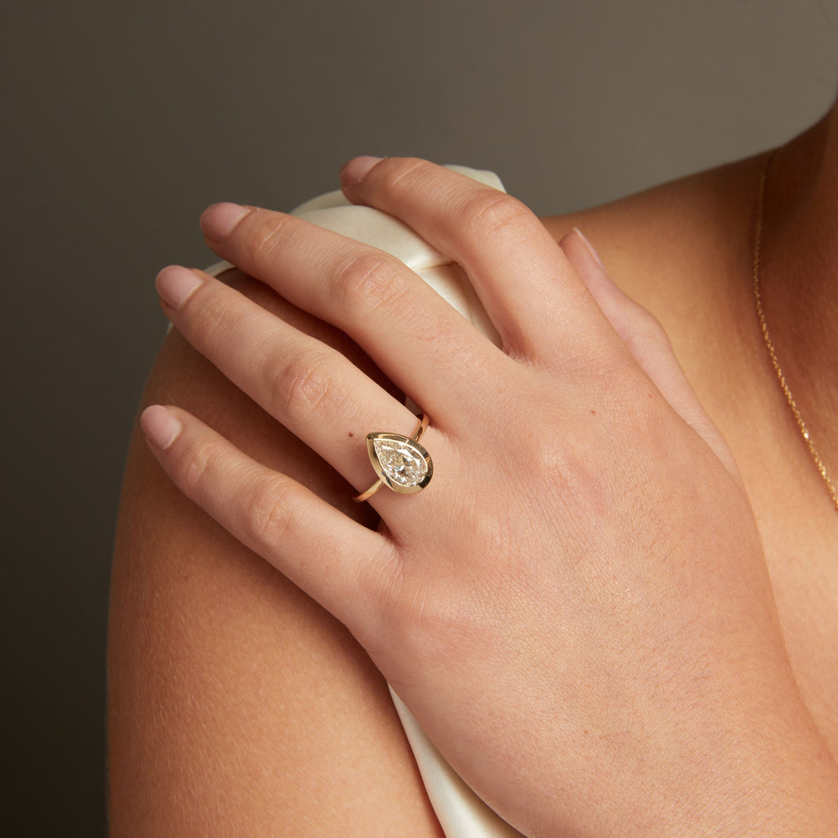 Model. A close-up lifestyle shot of a woman wearing a delicate gold ring with a pear diamond on her finger, posed against a soft neutral background.