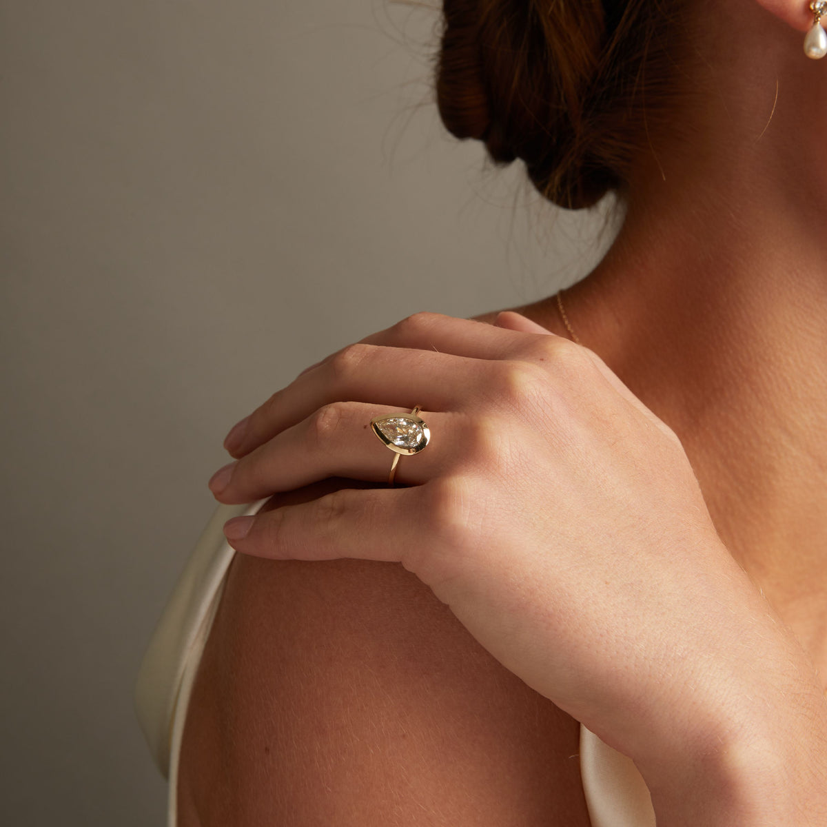 Model. A close-up lifestyle shot of a woman wearing a delicate gold ring with a pear diamond on her finger, posed against a soft neutral background.
