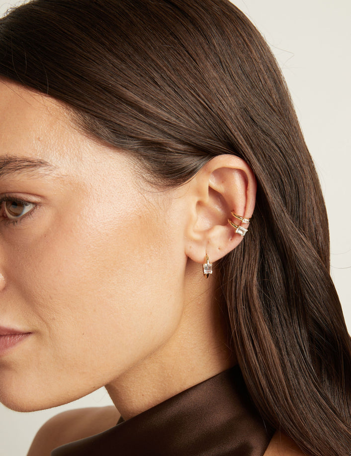 Model. Close up of woman wearing two gold ear cuffs and a huggie earring with white sapphires on neutral background. 