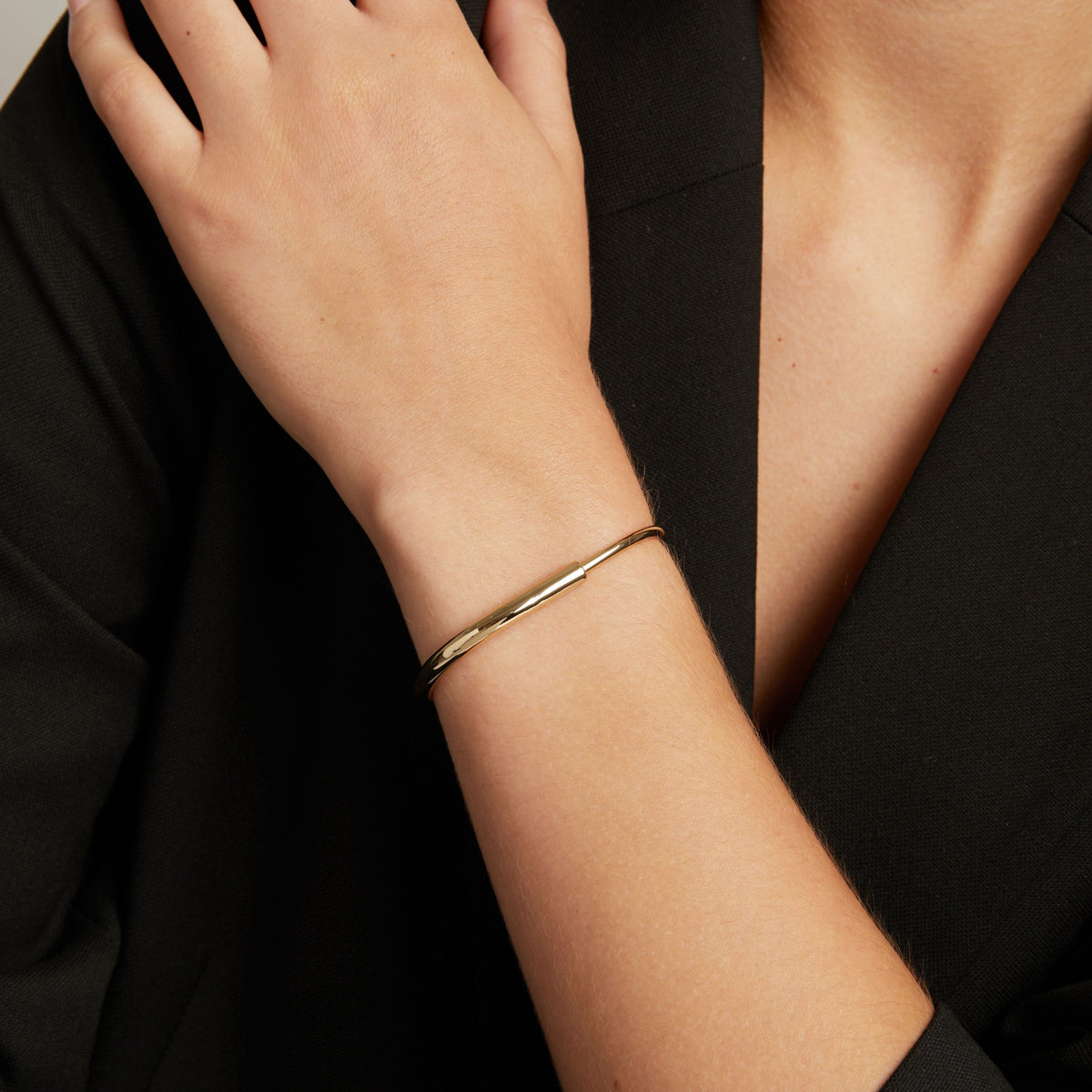 A close-up of a person's wrist wearing a thin, polished gold bangle bracelet, dressed in a black top.
