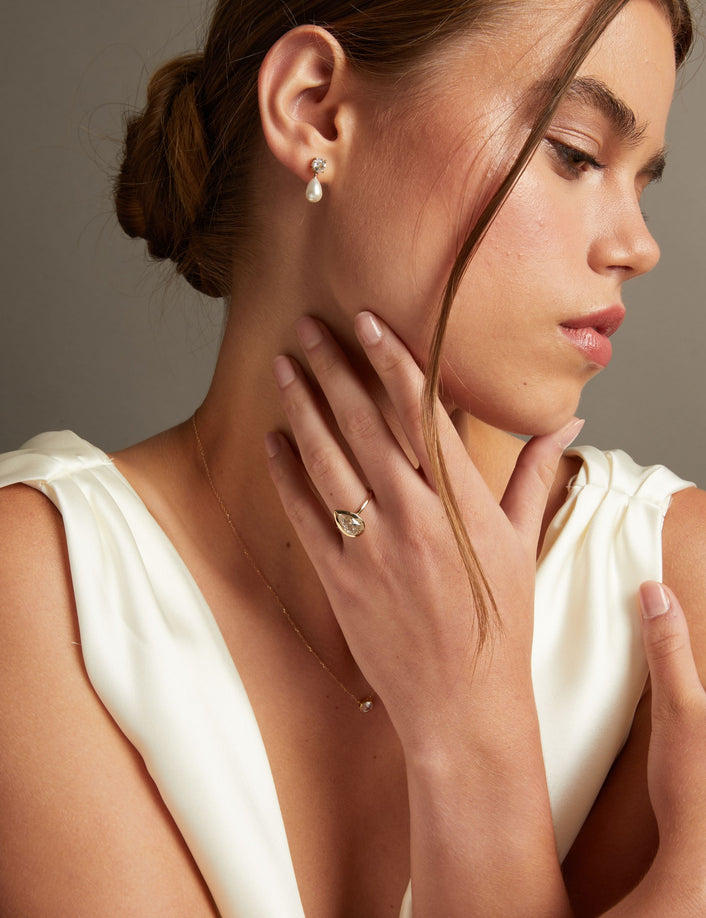 Model. A close-up portrait of a woman wearing delicate jewelry, including a small gold ring with a pear diamond on her finger and a matching pear drop earring on her ear. She is wearing a white satin dress against a neutral gray background.