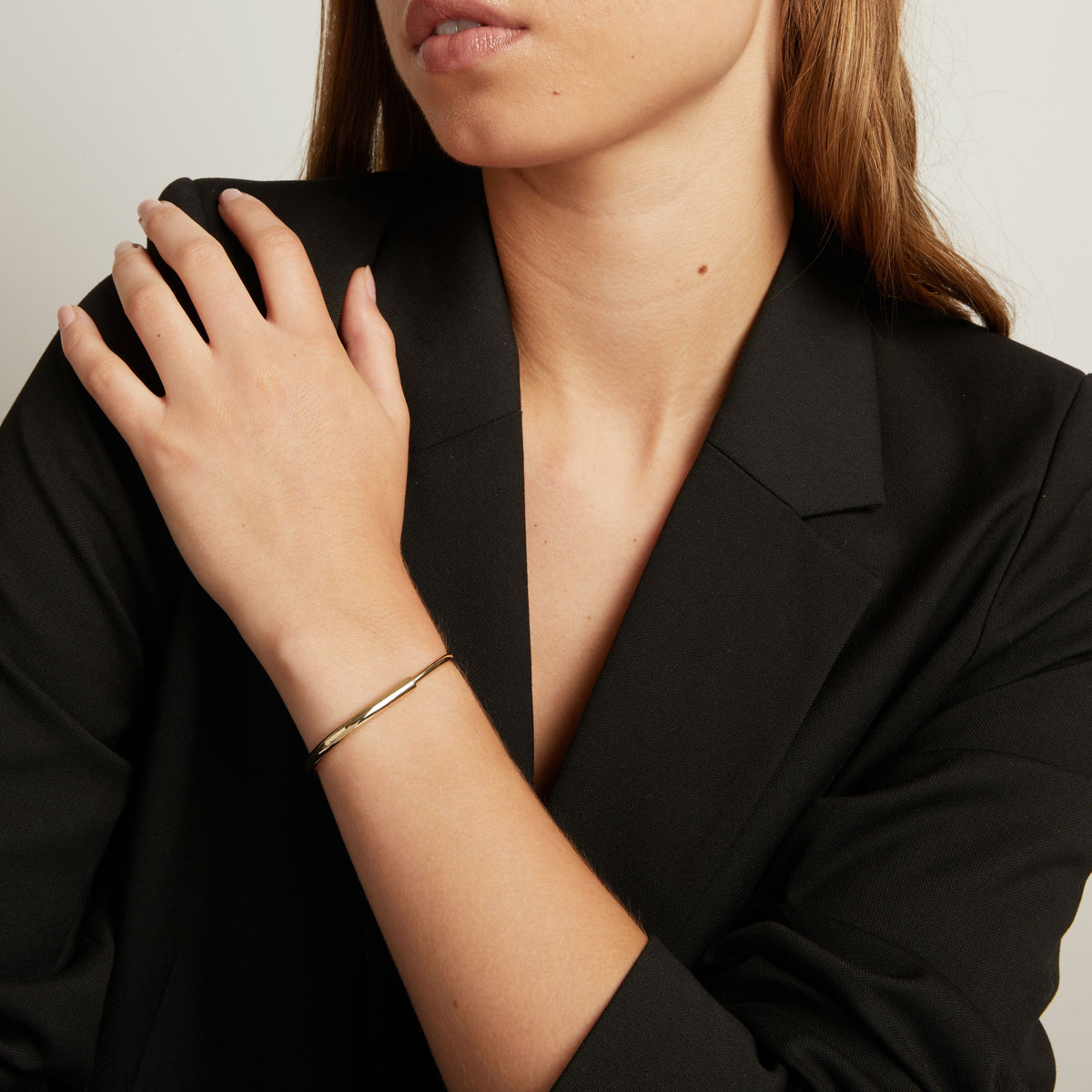 Model. A close-up of a woman wearing a slim, polished gold bangle bracelet on her wrist, paired with a black blazer against a light gray background.
