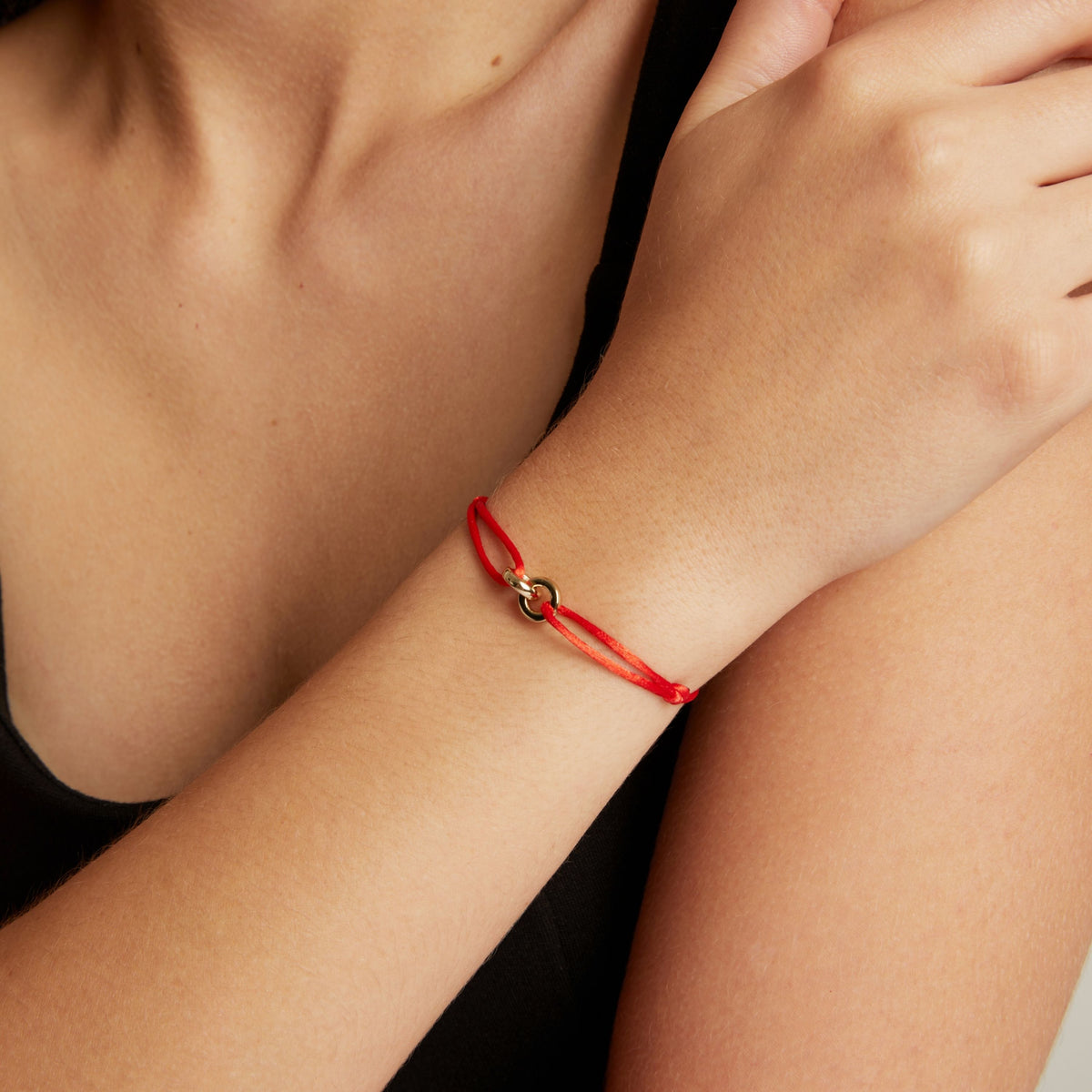 Model. A close-up of a person's wrist wearing a delicate cord bracelet with a small gold-toned interlocking link charm.