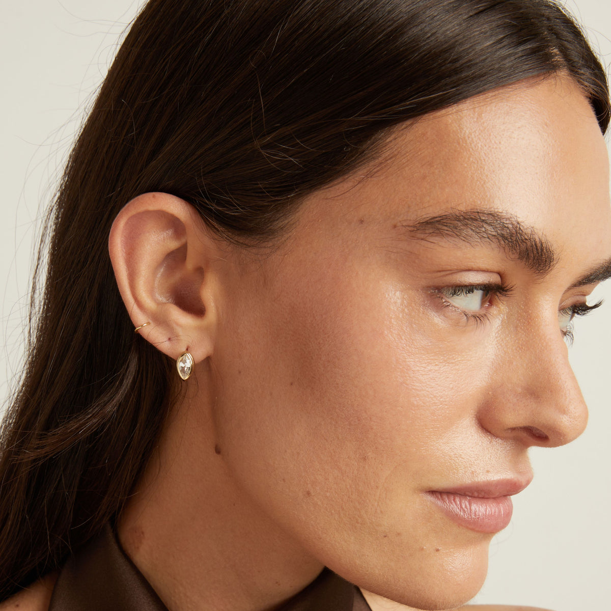 Close up of woman wearing a gold bezel huggie earring on neutral background.