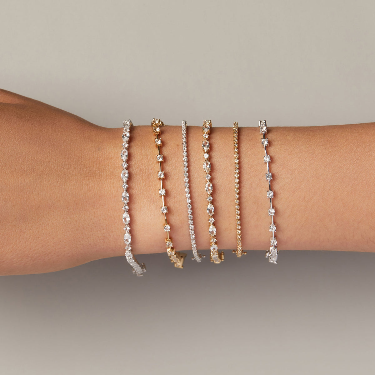 Model. Five delicate bangle bracelets stacked on a person's wrist, featuring silver, yellow gold, and white gold designs with clear pavé and star-set stones, against a neutral gray background.