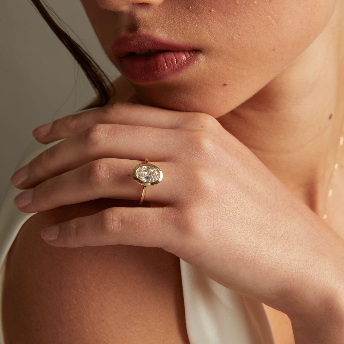 Model. A close-up lifestyle shot of a woman wearing a delicate gold ring with an oval diamond on her finger, posed against a soft neutral background.