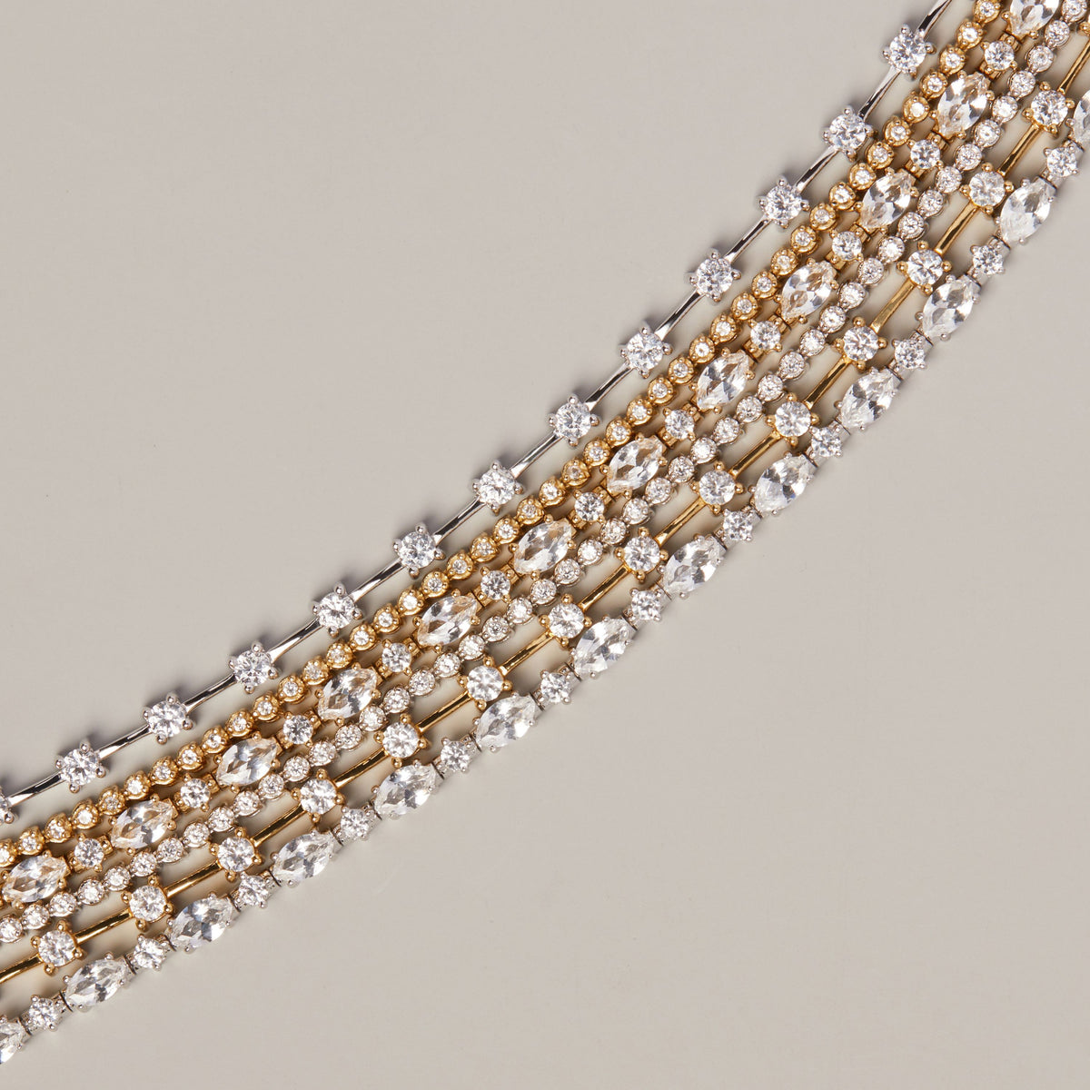Model. Close-up of a multiple diamond necklaces with gold and silver-toned metal settings, arranged diagonally on a neutral beige background.