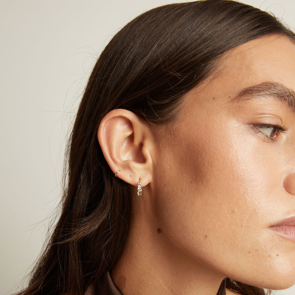 Close up of woman wearing a white sapphire huggie earring on a neutral background.