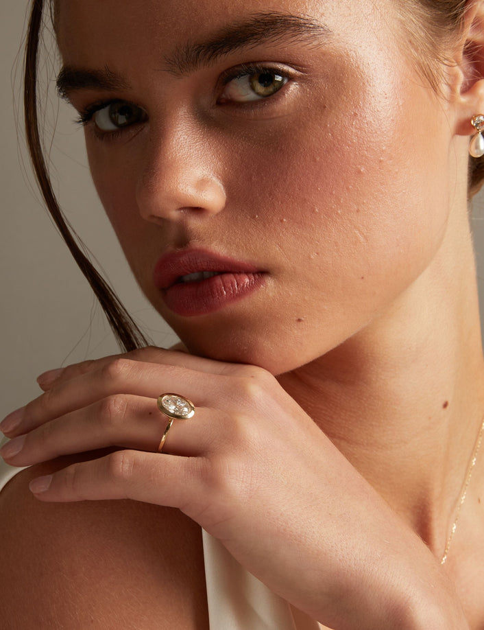 Model. A close-up lifestyle shot of a woman wearing a delicate gold ring with an oval diamond on her finger, posed against a soft neutral background.