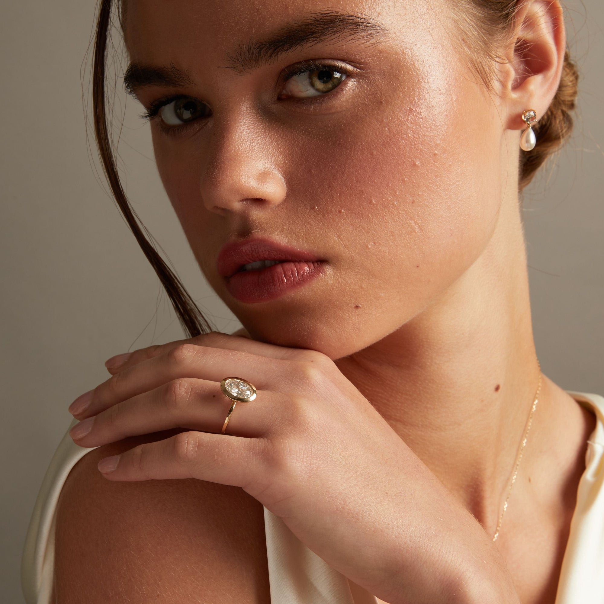Model. A close-up lifestyle shot of a woman wearing a delicate gold ring with an oval diamond on her finger, posed against a soft neutral background.