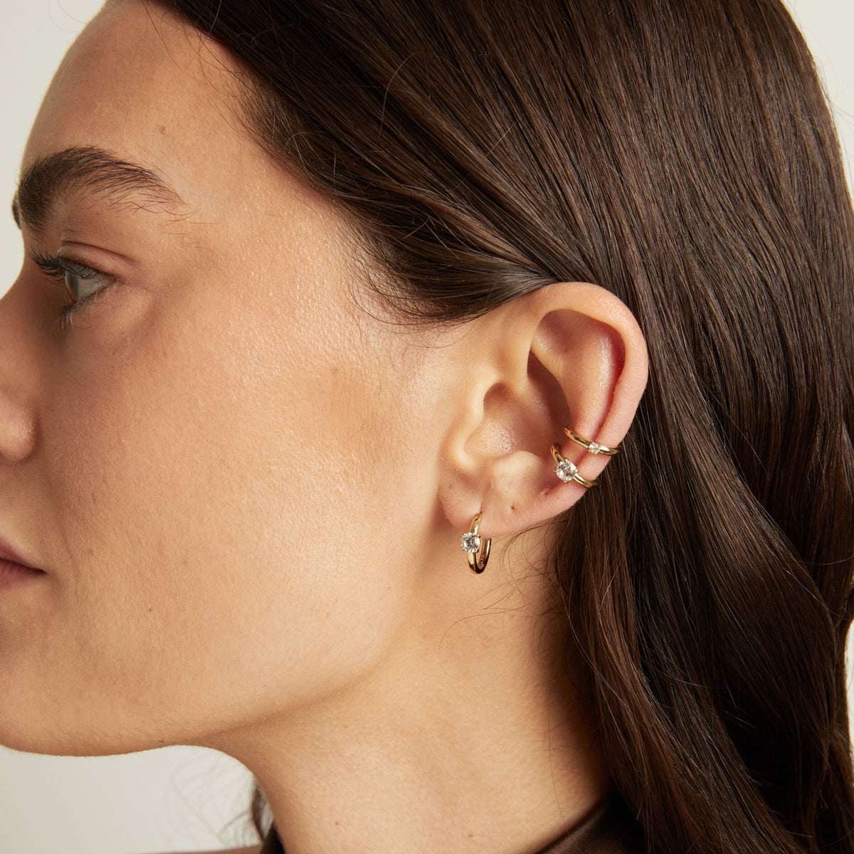 Close up of woman wearing two gold ear cuffs and a huggie earring with white sapphires on neutral background. 