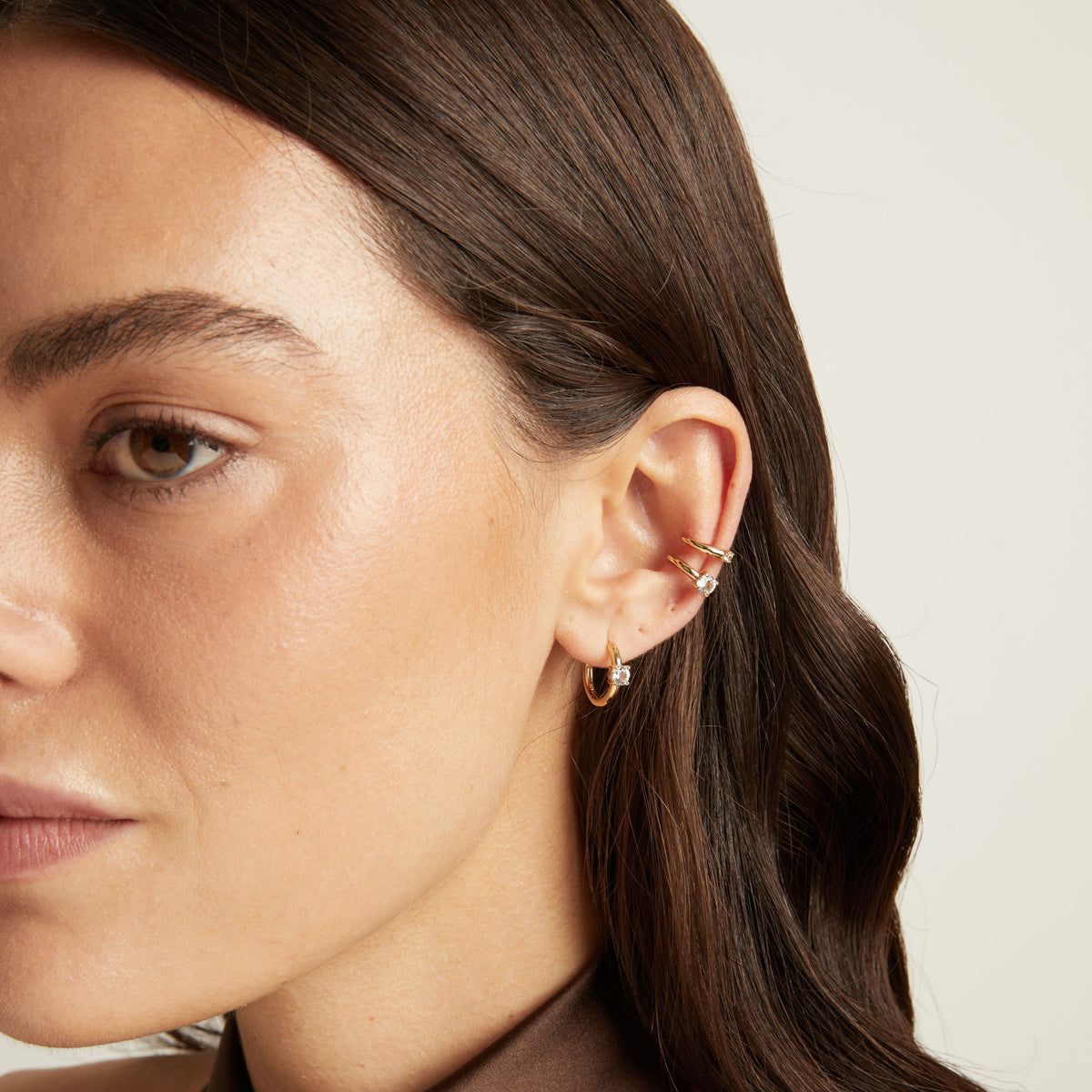 Model. Close up of woman wearing two gold ear cuffs and a huggie earring with white sapphires on neutral background. 