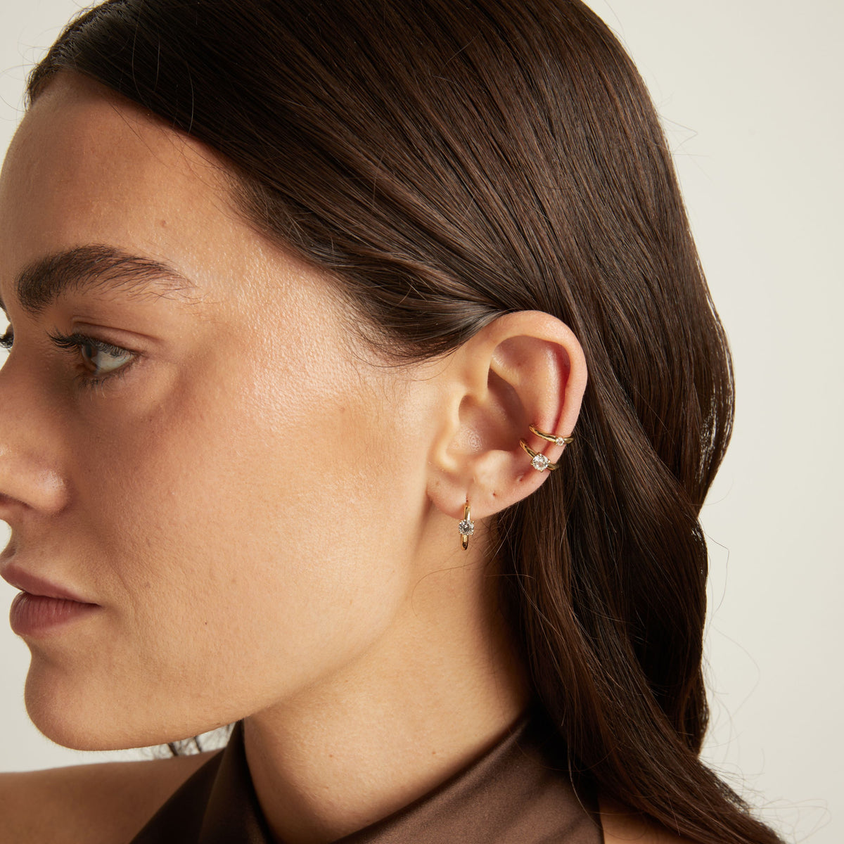 Close up of woman wearing two gold ear cuffs and a huggie earring with white sapphires on neutral background. 