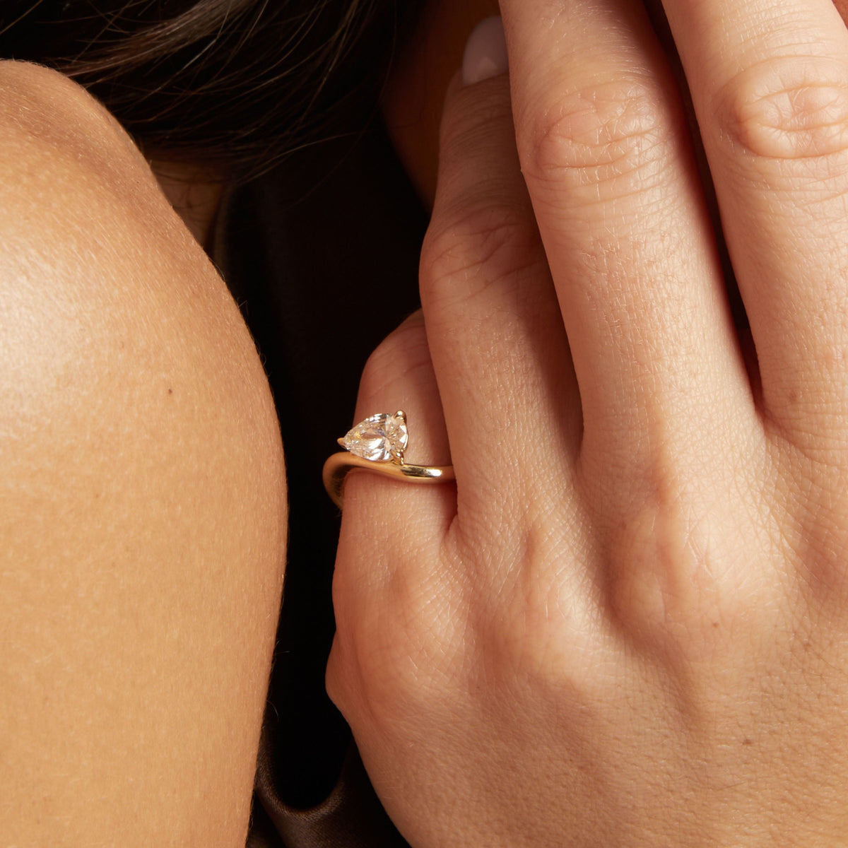 A close-up lifestyle shot of a woman wearing a gold ring with a floating pear diamond. 