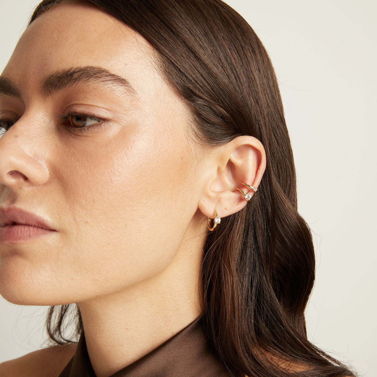 Close up of woman wearing two gold ear cuffs and a huggie earring with white sapphires on neutral background. 