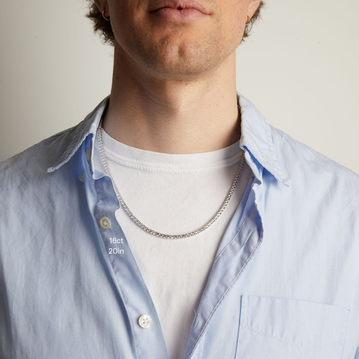 Model. A close-up of a man wearing a tennis necklace over a white t-shirt and a light blue button-down shirt.