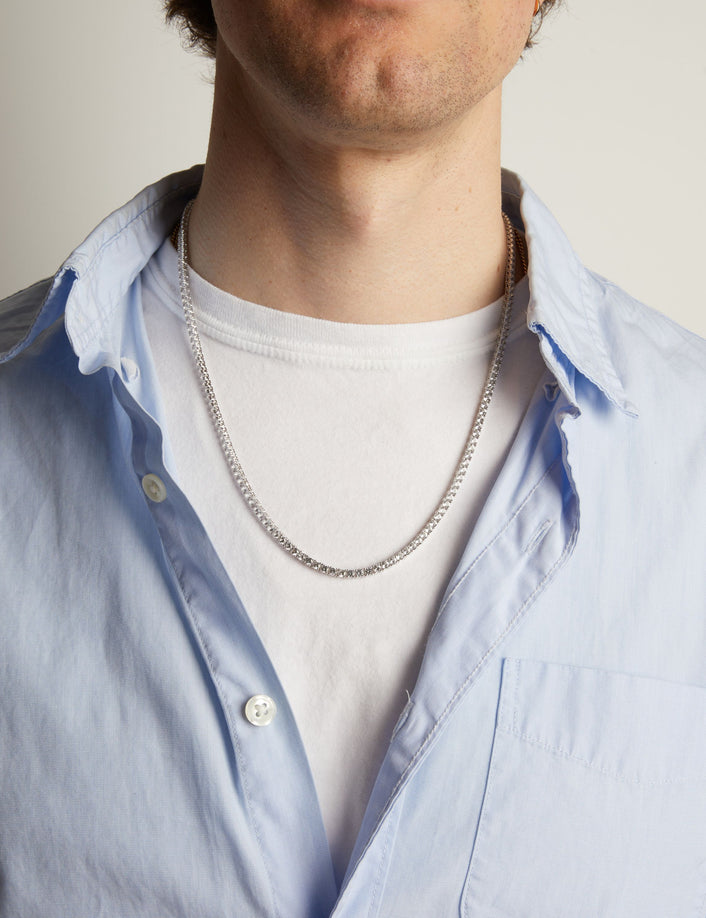 A close-up of a man wearing a tennis necklace over a white t-shirt and a light blue button-down shirt.